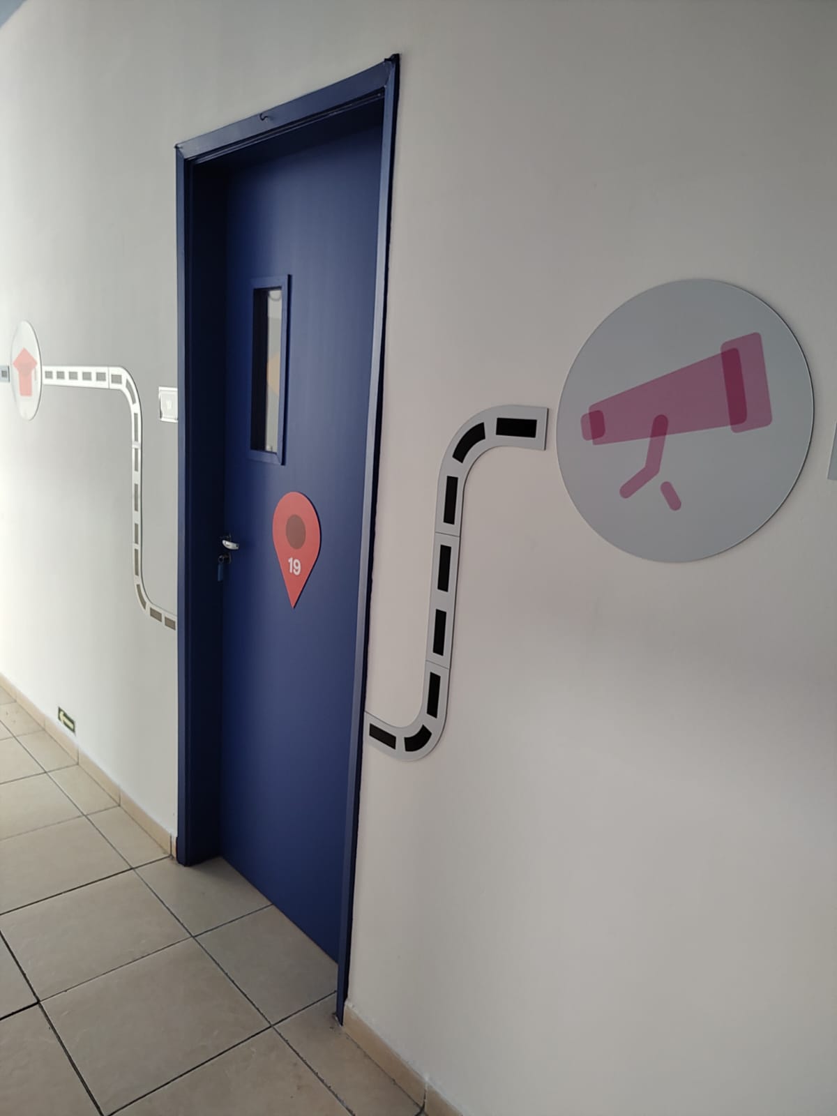 Corredor da escola com porta azul identificada pelo número 19 e placa circular com ilustração