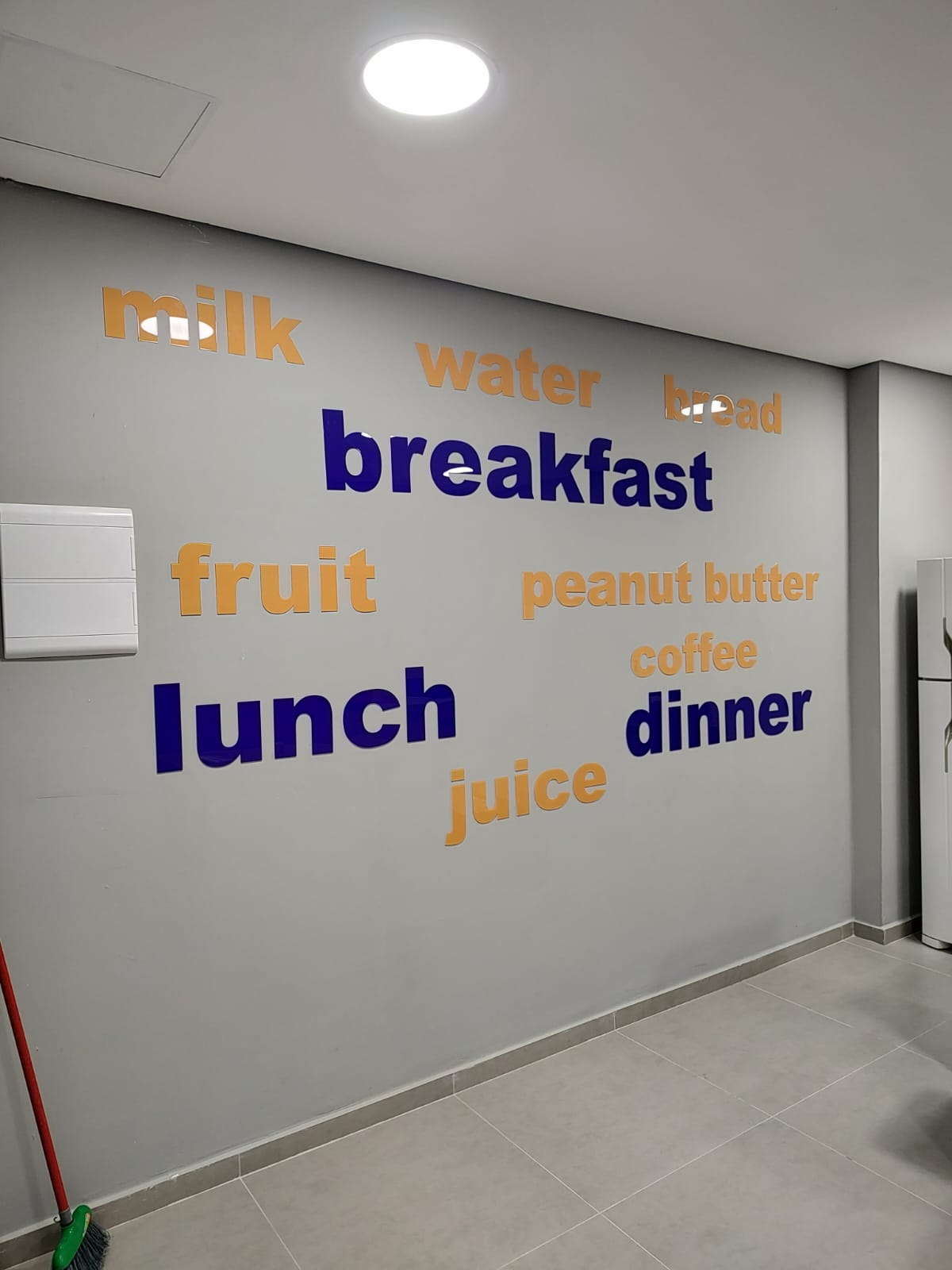 Sala de aula com palavras em azul e amarelo aplicadas na parede, como breakfast, lunch, dinner e fruit