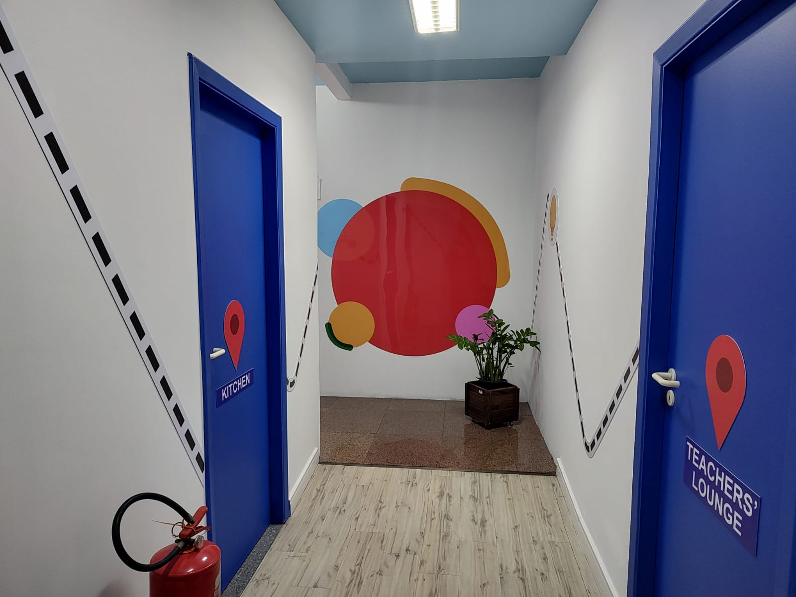 Corredor com portas azuis identificadas como Kitchen e Teachers’ Lounge e painel circular colorido ao fundo